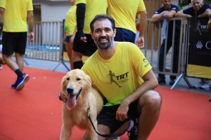 Na imagem, há um homem ajoelhado ao lado de um cachorro, ambos na frente de uma área delimitada por grades de metal para um evento de corrida. O homem está sorrindo e veste uma camiseta amarela com o logotipo "TRT" e o desenho de um corredor, além de um par de shorts escuros e tênis esportivos. Ele tem barba escura e cabelo curto, e aparenta estar relaxado e feliz.O cachorro é um Golden Retriever, com uma expressão amigável, a língua para fora, sugerindo que ele pode ter estado ativo ou estar com calor. Ele está usando uma coleira e parece estar olhando para a câmera com um olhar atento e alegre.No fundo, outras pessoas estão usando roupas esportivas amarelas, sugerindo que todos são participantes do mesmo evento. O ambiente parece ser ao ar livre e informal, com um tapete vermelho no chão, indicando a área de corrida ou chegada.