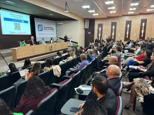 Imagem colorida mostrando diversas pessoas reunidas em um auditório. Há uma plateia, sentada nas cadeiras do auditório. Na frente da plateia há um palco, onde algumas pessoas estão sentadas atrás de um balcão retangular de madeira. Atrás das pessoas no palco, há um telão branco com uma projeção.
