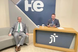 Foto de um homem sentado em uma poltrona e um homem numa bancada. Eles olham para a câmara. Ao fundo, a logomarca do programa hora extra. Embaixo da bancada, a logomarca da Justiça do Trabalho