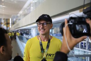 Na imagem, vemos um homem sorridente vestindo uma camiseta amarela, boné preto e uma credencial pendurada no pescoço. Ele parece estar em um ambiente interno iluminado e espaçoso, um centro de convenções, dado o visual moderno e a presença de corrimões como de escadas rolantes ou balcões de atendimento ao fundo. A direita da imagem, uma pessoa está segurando uma câmera de vídeo, sugerindo que o homem de camiseta amarela está sendo filmado ou fotografado. Ele parece estar em um bom humor e está em um evento, como indicado pela credencial.