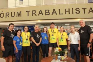 A imagem mostra um grupo de oito pessoas posando para uma fotografia no fórum trabalhista, como indicado pela placa acima deles. As pessoas estão vestidas em diferentes cores de camisetas, algumas das quais exibem o logotipo "TRT" semelhante ao visto nas outras imagens, sugerindo que elas participaram de um evento esportivo relacionado ao Tribunal Regional do Trabalho.Algumas pessoas estão usando medalhas, o que implica que elas completaram a corrida. As cores das camisetas variam de amarelo a azul e preto, com algumas camisetas mostrando números de corrida. O grupo parece estar em um espaço de recepção ou área de espera, com mesas e flores decorativas, sugerindo que o local pode ser utilizado para eventos ou celebrações.Todos estão sorrindo e parecem estar satisfeitos, indicando um ambiente de celebração de um evento pós-corrida.