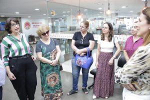 A imagem mostra um grupo de mulheres em um ambiente interno. Duas mulheres estão no centro da imagem conversando ou ouvindo atentamente; uma delas está gesticulando como se estivesse falando. As outras mulheres estão olhando em direção à pessoa que está falando, mostrando interesse no que está sendo dito. A iluminação é interna e artificial, e o ambiente parece ser casual e informal, com as pessoas vestidas confortavelmente para a ocasião.