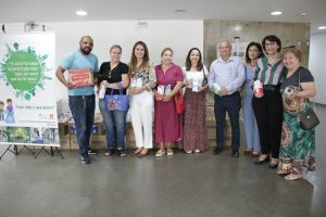 A imagem mostra um grupo de nove pessoas em um ambiente interno. Elas estão de pé, alinhadas lado a lado, e participam de um evento de doação, como sugerido pelo banner ao fundo com uma mensagem sobre a importância de contribuir independentemente do tamanho da ação. Algumas pessoas estão segurando caixas de leite, simbolizando as doações mencionadas no banner. O ambiente tem um piso claro e paredes de concreto aparente. A presença de caixas empilhadas ao fundo sugere que mais itens aguardam distribuição. Todos os presentes exibem expressões contentes.