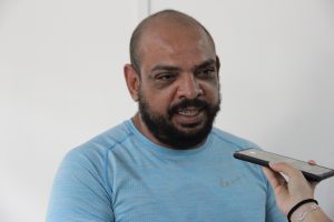 A imagem mostra um homem uma camisa azul e olha para um dispositivo móvel que está sendo segurado por outra pessoa fora da cena, indicando que ele pode estar dando uma entrevista ou respondendo a perguntas. O fundo é uma parede branca, e a cena parece capturada em um contexto de trabalho ou evento social.