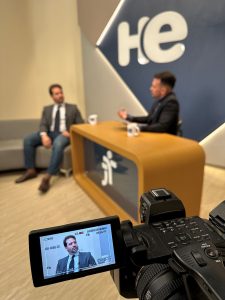 Dois homens no estúdio do programa Hora Extra. Um sentado em um sofá cinza. O outro está sentado atrás de uma bancada que tem na frente a logo do TRT. Atrás há um painel com a logo do programa Hora Extra. Em cima da bancada, duas canecas com a logo do Hora Extra. No canto esquerdo inferior está a câmara de tv e no visor a imagem do entrevistado, que está sentado no sofá