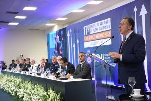 Foto colorida do ministro Luís Roberto Barroso durante o 17º ENPJ. Trata-se de um homem branco, de cabelos grisalhos. Ele usa um terno preto e está atrás de um púlpito falando ao microfone. Ao fundo, nove pessoas estão sentadas atrás de uma mesa, na frente do painel de divulgação do 17º ENPJ.