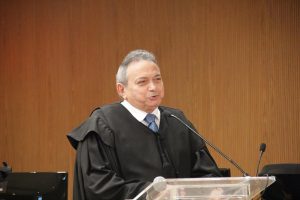 Foto do desembargador Geraldo Nascimento discursando. Trata-se de um homem branco, de cabelo curto, liso e branco. O magistrado veste um terno sobreposto por uma toga. Ele está de pé atrás de um púlpito falando no microfone de mesa.