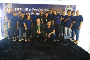 A imagem mostra um grupo de pessoas reunidas para uma foto. Há um total de dezesseis pessoas, algumas em pé e outras agachadas na frente. O grupo parece ser uma mistura de jovens e adultos vestindo camisetas azuis, com exceção de um homem no centro, que está agachado e veste um terno claro e uma gravata. Eles estão posando em frente a um fundo que inclui o texto "TRT-18 e Programa Trabalho Seguro", sugerindo que pode ser uma iniciativa relacionada ao Tribunal Regional do Trabalho da 18ª Região no Brasil. A imagem tem uma atmosfera formal, mas amigável.