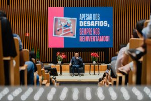 Foto feita do fundo do auditório, mostra de forma desfocada as pessoas sentadas e, no centro, sobre o palco, o palestrante sentado em uma poltrona segurando o microfone, ao fundo, o telão de LED exibe os dizeres "apesar dos desafios, sempre nos reinventamos"