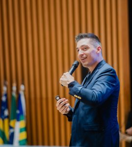 Foto de Samuel Bortolin. Trata-se de um homem branco de cabelo curto liso grisalho. Ele veste um terno preto e fala em um microfone de mão.