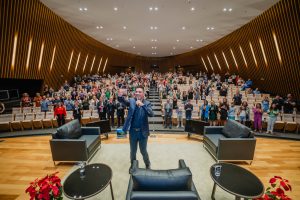 Foto do palestrante e plateia.  O palestrante está de pé, no centro do palco, e segura o celular para uma "selfie", ao fundo, o público espectador do auditório, também de pé, pousa para a foto.