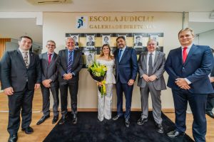 Foto da desembargadora Iara, no centro, em pé, segurando um buquê de flores, está com roupa social branca, ao lado dela estão seis homens em pé de terno e gravata