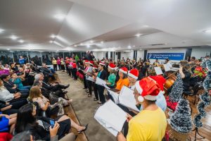 Foto colorida mostra um grupo de pessoas se apresentando para uma plateia. Ao fundo, uma orquestra tocando. O grupo de pessoas usa blusa de diferentes cores e um gorro vermelho com detalhes brancos na cabeça