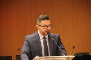 Foto do Procurador Tiago Ranieri discursando. Trata-se de um homem pardo, de cabelo curto, liso e grisalho. Ele veste um blazer cinza escuro, camisa branca, gravata preta e usa óculos de armação preta. Ele está de pé atrás de um púlpito falando em um microfone de mesa.