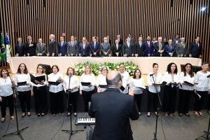 A imagem mostra um evento formal em um ambiente interno com decoração elegante. No primeiro plano, há um coro de adultos mistos, com cerca de 15 membros, todos vestidos de preto e branco. Os cantores, possivelmente sopranos, altos, tenores e baixos, estão alinhados em uma formação de coro, segurando pastas de partituras e focados em um maestro que está de costas para a câmera, conduzindo-os com a mão esquerda erguida. Atrás do coro, há um grupo de homens vestidos com ternos formais e gravatas, em pé e alinhados em uma única fileira. Eles estão em frente a uma parede de madeira com um design vertical e a bandeira do Brasil ao centro, indicando que o evento pode ser no Brasil. A iluminação é uniforme e parece ser interna, realçando o caráter formal do evento.