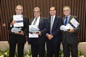 A imagem mostra quatro homens vestidos com trajes formais, de pé e alinhados lado a lado, segurando certificados e prêmios. Eles estão sorrindo e olhando diretamente para a câmera, indicando que esta é uma foto posada. Cada um deles segura um documento com uma capa branca e azul. O cenário tem uma parede de madeira vertical e uma decoração composta por folhagens e flores, sugerindo que a foto foi tirada em um evento formal, possivelmente uma cerimônia de premiação ou reconhecimento. A iluminação da sala é suave e uniforme.