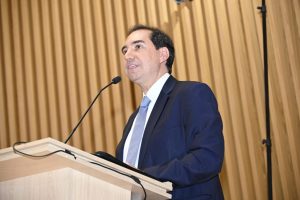 A imagem mostra um homem vestido formalmente falando em um púlpito. Ele usa um terno escuro, camisa branca e gravata. O homem parece estar falando ativamente, possivelmente em um discurso ou apresentação, considerando sua expressão facial e boca aberta. O púlpito é de cor clara e simples, e há um microfone ajustado à sua altura. Ao fundo, podemos ver uma parede com um design vertical de ripas de madeira.