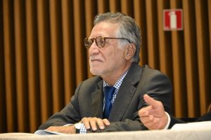 A foto apresenta um homem de cabelos brancos sentado, parecendo estar em uma reunião ou conferência. Ele está vestindo um terno escuro com uma camisa de colarinho aberto e uma gravata com padrão de xadrez. O foco está no homem, com o fundo levemente desfocado, mas ainda é possível identificar a parede de ripas de madeira ao fundo. A iluminação é suave e centrada no sujeito.