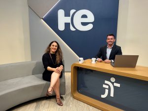 Uma mulher sentada numa poltrona e um homem numa bancada. Atrás, na parede, a logo do programa Hora Extra HE.