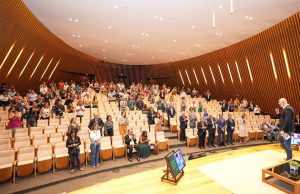 Foto do auditório com pessoas sentadas nas poltronas e o palestrante Tande no palco