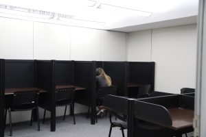 A imagem é de uma sala de estudos com uma atmosfera mais reservada. Há cabines de estudo individuais alinhadas, cada uma com uma cadeira e uma mesa pequena. Uma pessoa com cabelo loiro está sentada em uma das cabines, de costas para a câmera, focada em seu trabalho. A iluminação é suave e o espaço parece ser destinado a estudo ou trabalho que requer concentração.