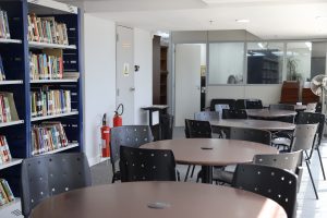 A imagem mostra uma biblioteca ou sala de estudos bem iluminada e espaçosa. Há várias mesas redondas com cadeiras pretas ao redor, prontas para os usuários. Uma estante de livros cheia se destaca em primeiro plano à esquerda, e no fundo há uma janela de vidro que revela outra sala com uma pessoa sentada. A cena transmite um ambiente tranquilo para leitura ou estudo.