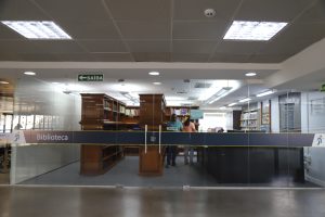 Na imagem vemos a entrada de uma biblioteca, marcada por uma barreira baixa com a palavra "Biblioteca" e uma seta indicando a saída. Pode-se ver um balcão de atendimento com duas pessoas conversando e mais estantes de livros ao fundo. A iluminação é clara e a área parece bem organizada, indicando a entrada para um ambiente de estudo e leitura.


