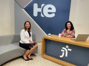 Uma mulher sentada em uma poltrona e uma mulher numa bancada. Elas olham para a câmara. Ao fundo, a logomarca do programa hora extra. Embaixo da bancada, a logomarca da Justiça do Trabalho. É um estúdio de TV