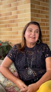 Mulher branca, de cabelos castanhos na altura do ombro, vestindo blusa preta e calças amarelas. Elas está sentada e ao fundo há uma parede de tijolinho amarelo