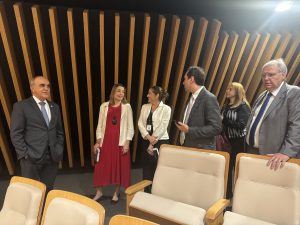 Algumas pessoas em é conversando. À frente cadeiras de um auditório. Atrás, uma parede com detalhes em madeira