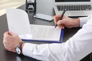 Na imagem um homem está analisando um documento em cima de uma mesa com um notebook ao fundo