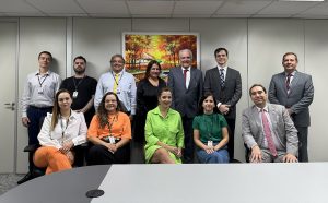 Foto mostra o corregedor e outros servidores em pé contra uma parede na qual há um quadro. À frente deles estão outros servidores  e um juiz sentados em cadeiras. 