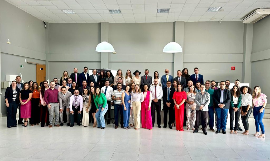 Dezenas de pessoas posando para a foto em um ambiente fechado com duas luminárias ao centro