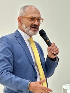 Homem de terno azul e gravata amarela falando ao microfone