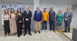 Foto colorida mostra nove pessoas, entre homens e mulheres vestidos em trajes formais em uma ambiente fechado e claro. Eles estão posando para a foto e logo atrás deles há uma parede.
