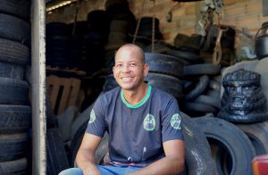 Foto do artista plástico Cristiano Borges. Ele é um homem calvo, de pele morena, usa calça jeans e camiseta e está sentado sorrindo para a câmera. No fundo, pneus usados amontoados no ambiente de trabalho dele.