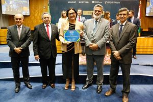 Foto mostra cinco pessoas, sendo quatro homens e uma mulher, lado a lado e posando para a foto. A mulher, desembargadora Wanda Ramos, está no meio e segura o certificado recebido na premiação 