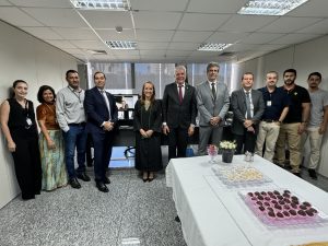 Onze pessoas estão posicionadas para uma foto em um escritório. São três mulheres e oito homens, todos vestidos de maneira formal. A decoração é simples, com foco nos participantes e na mesa de doces à frente deles.