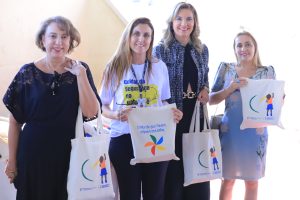 A imagem mostra quatro mulheres sorrindo e posando juntas em um ambiente claro, possivelmente um evento. Elas seguram sacolas que parecem ter logotipos de organizações ou campanhas, indicando que podem estar em um evento corporativo ou de caridade. Da esquerda para a direita: a primeira mulher usa uma blusa de renda azul, a segunda veste uma camiseta com o que parece ser um logotipo de evento, a terceira está em um traje formal de blazer padronizado, e a quarta veste uma vestido. Todas as mulheres parecem estar envolvidas ou participando do evento representado nas sacolas que seguram.