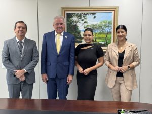 A imagem mostra quatro pessoas posando para uma fotografia em um ambiente de escritório formal. Eles estão de pé diante de uma mesa de madeira escura e atrás deles há uma pintura grande de uma paisagem rural emoldurada em dourado. Todos estão com uma expressão amigável e profissional, indicando que podem ser colegas ou participantes de um evento profissional.
