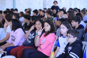 A imagem mostra um grupo de estudantes adolescentes sentados em um amplo local aberto, assistindo a uma apresentação. As expressões e posturas variam: alguns alunos estão atentos, outros conversando discretamente, e outros posam para a foto.
