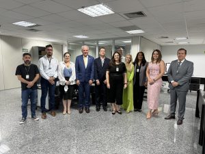 Na imagem, vemos um grupo de pessoas posando para uma fotografia em um ambiente de escritório. O local parece ser um escritório amplo, bem iluminado, com paredes brancas e várias divisórias ao fundo que indicam outras áreas de trabalho. O piso é de granito cinza.
Todas as pessoas estão vestidas formalmente. Eles estão alinhados lado a lado, todos sorrindo para a câmera, com crachás visíveis.