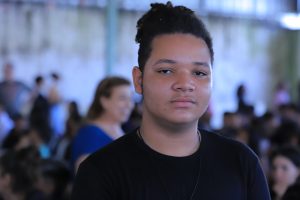 Foto de estudante em primeiro plano e, ao fundo, os outros formando uma fila.