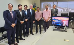 Nesta foto, sete homens estão posando em um escritório. Eles estão de pé, alinhados e sorrindo para a câmera. Todos vestem trajes formais: cinco deles estão de terno e gravata, enquanto os outros dois vestem camisas mais casuais. Ao fundo, é visível uma mesa de escritório com um computador mostrando uma paisagem de praia no monitor.