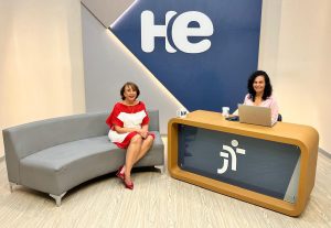 Uma mulher sentada em uma poltrona e um mulher numa bancada. Elas olham para a câmara. Ao fundo, a logomarca do programa hora extra. Embaixo da bancada, a logomarca da Justiça do Trabalho