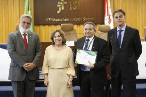 A imagem mostra quatro pessoas em pé, posando para uma foto em um ambiente formal, provavelmente durante um evento ou cerimônia, diante de um fundo com o logotipo do Tribunal Superior do Trabalho (TST) e a frase "O Tribunal da Justiça Social". Da esquerda para a direita, o primeiro homem veste um terno cinza com gravata vermelha e sorri, a segunda pessoa é uma mulher usando um vestido bege claro, também sorridente, o terceiro homem segura um certificado nas mãos e veste um terno preto com gravata azul, e o quarto homem está de terno escuro e gravata azul. Ao fundo, há uma decoração de madeira e parte da bandeira do Brasil é visível.