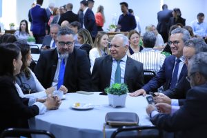A imagem mostra um grupo de pessoas reunidas em torno de uma mesa redonda em um evento formal. Três homens em trajes formais estão conversando no centro, cercados por outras pessoas que também interagem. A mesa possui plantas decorativas e itens como xícaras de café. Ao fundo, outras pessoas participam do evento em um ambiente bem iluminado.