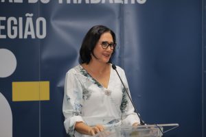 A imagem mostra uma mulher de meia-idade, com cabelos escuros e óculos, falando em um púlpito de acrílico transparente. Ela está usando uma blusa clara com estampas florais. Ao fundo, há um painel azul que exibe parte das palavras "REGIONAL DO TRABALHO" e "REGIÃO", sugerindo que o evento está relacionado a uma instituição de trabalho. A mulher sorri levemente, o que indica uma abordagem amigável e confiante durante sua fala.