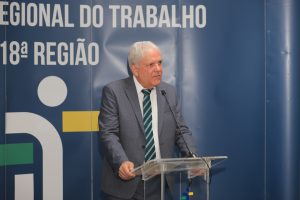 A imagem mostra um homem idoso em traje formal (terno cinza e gravata listrada) falando em um púlpito com microfone. Ele está em frente a um painel azul que exibe as palavras "REGIONAL DO TRABALHO 18ª REGIÃO", sugerindo que o evento está relacionado a uma instituição de trabalho ou justiça laboral. A expressão do homem é séria e atenta, indicando que ele está fazendo um discurso ou uma apresentação oficial.