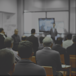 Esta imagem retrata uma situação de reunião ou apresentação em um ambiente profissional. A cena está focada na perspectiva de alguém que está sentado entre várias outras pessoas, todos voltados para a frente, onde um apresentador está de pé diante de um grande monitor.